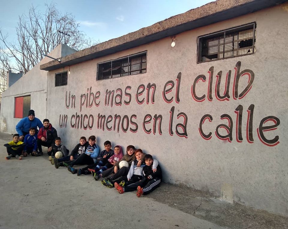 Chicos en un club con una pintada que dice "un pibe más en el club, un chico menos en la calle"